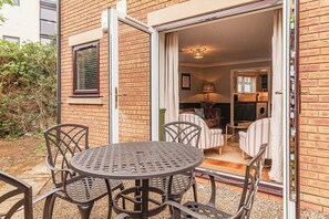 Een terras buiten met een ronde metalen tafel en stoelen, een glazen schuindeur naar een zithoek met een bank en een lamp, en een bakstenen muur.