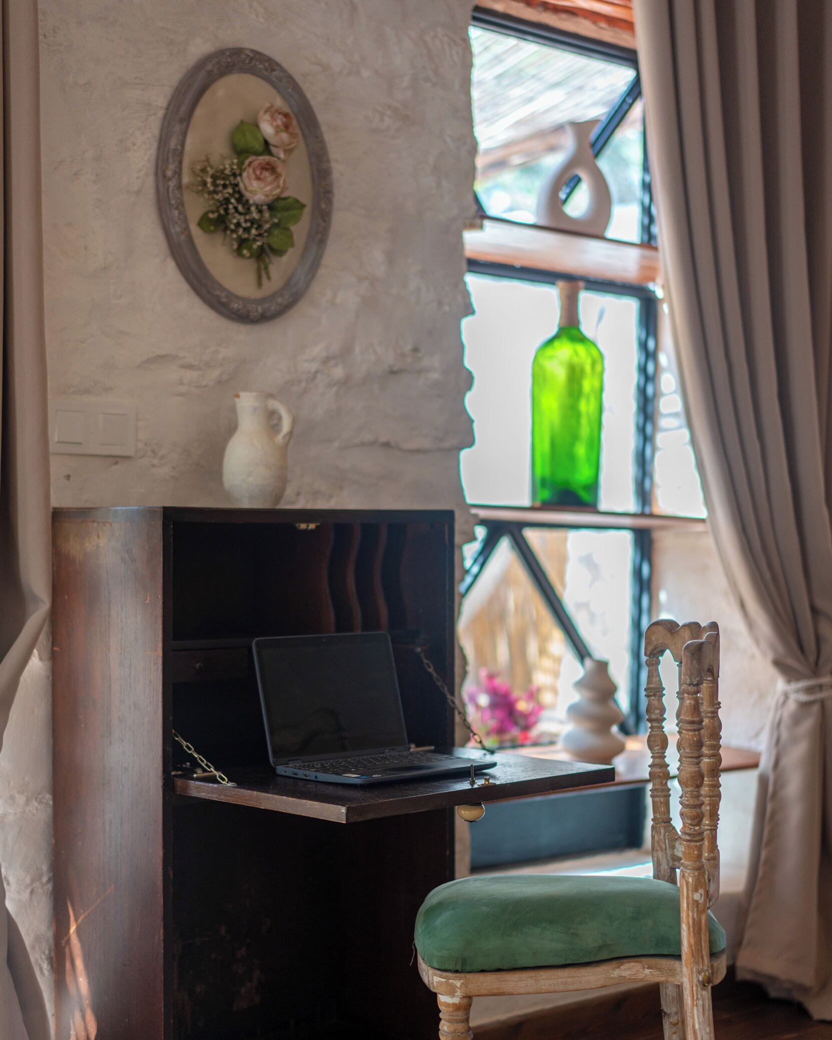 Una habitación con una computadora portátil sobre una estantería, una silla con un cojín verde, un espejo decorativo y una botella verde en una estantería.