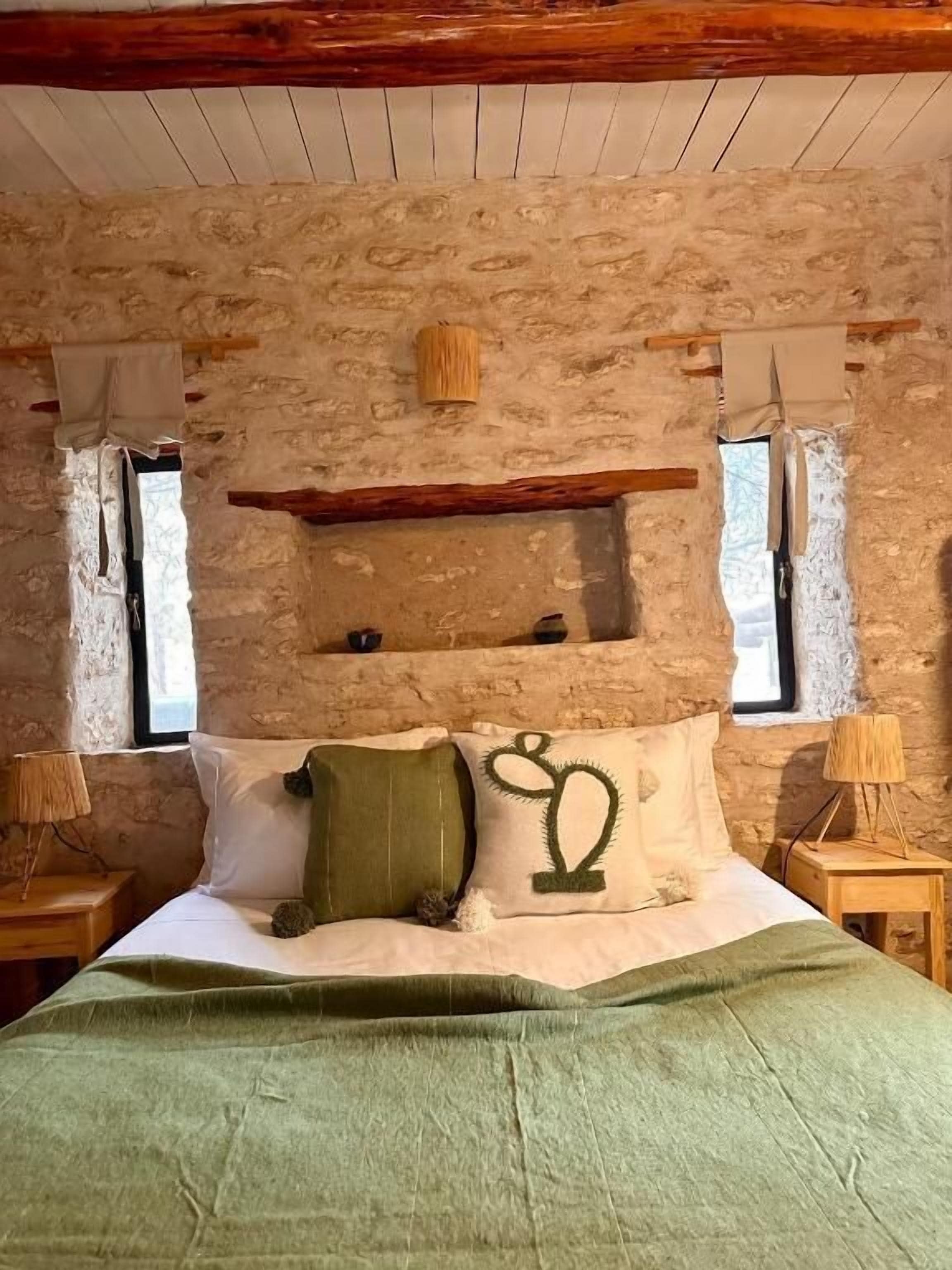 A rustic bedroom with a stone wall, wooden beams, a bed with green bedding, and two bedside lamps.