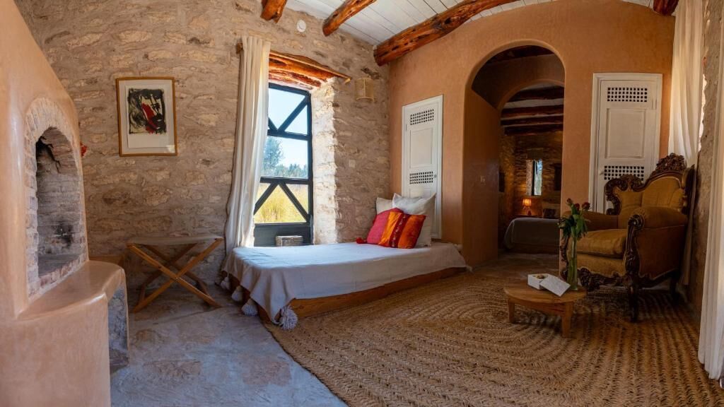 Una habitación rústica con una cama, una silla de madera, una mesita y un muro de piedra.