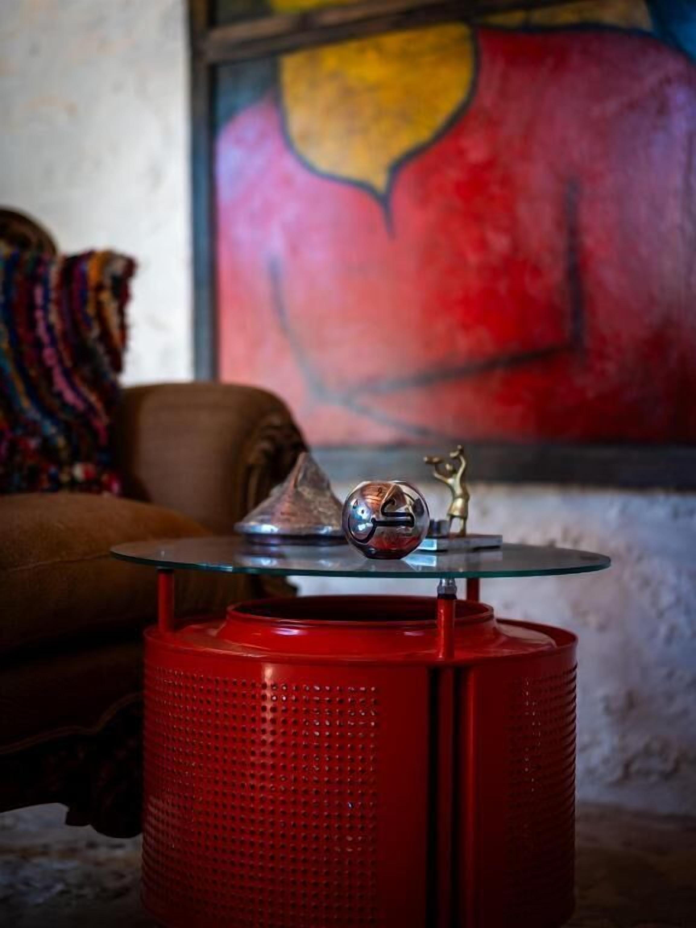 A red, perforated heater with a glass top and a metallic knob, placed on a wooden floor.