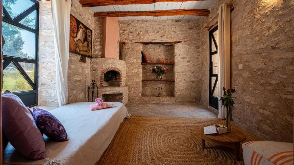A rustic room with a stone fireplace, a bed with pillows, a wooden coffee table, and a view through a window.