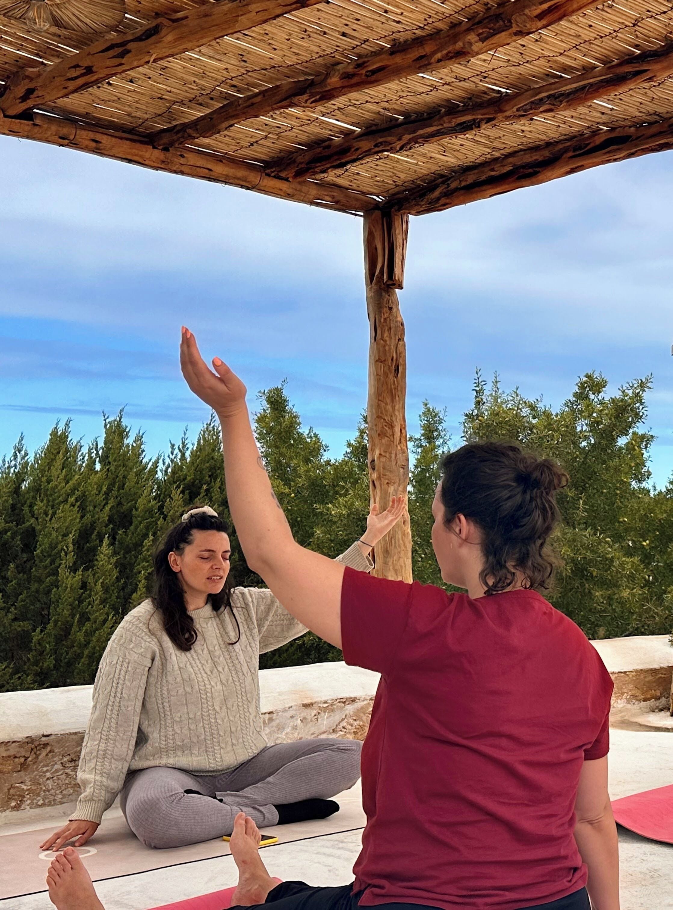 Two people sitting on a mat outdoors under a wooden structure with a clear sky.