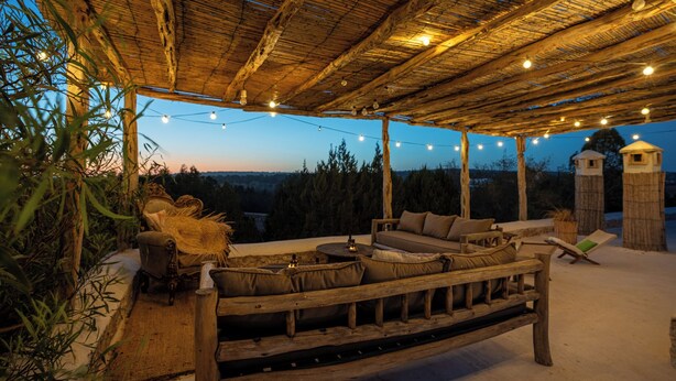 An outdoor seating area with a wooden bench and cushions, string lights, and a view of the forest. - Atlantis 12 Maison d’hôtes et d’art (Ounagha)