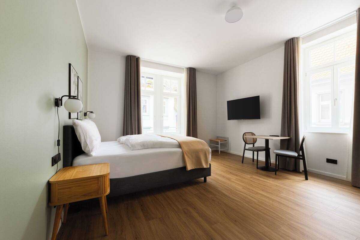 Una camera d'albergo moderna con un letto, un comodino, una TV e una scrivania con sedie.