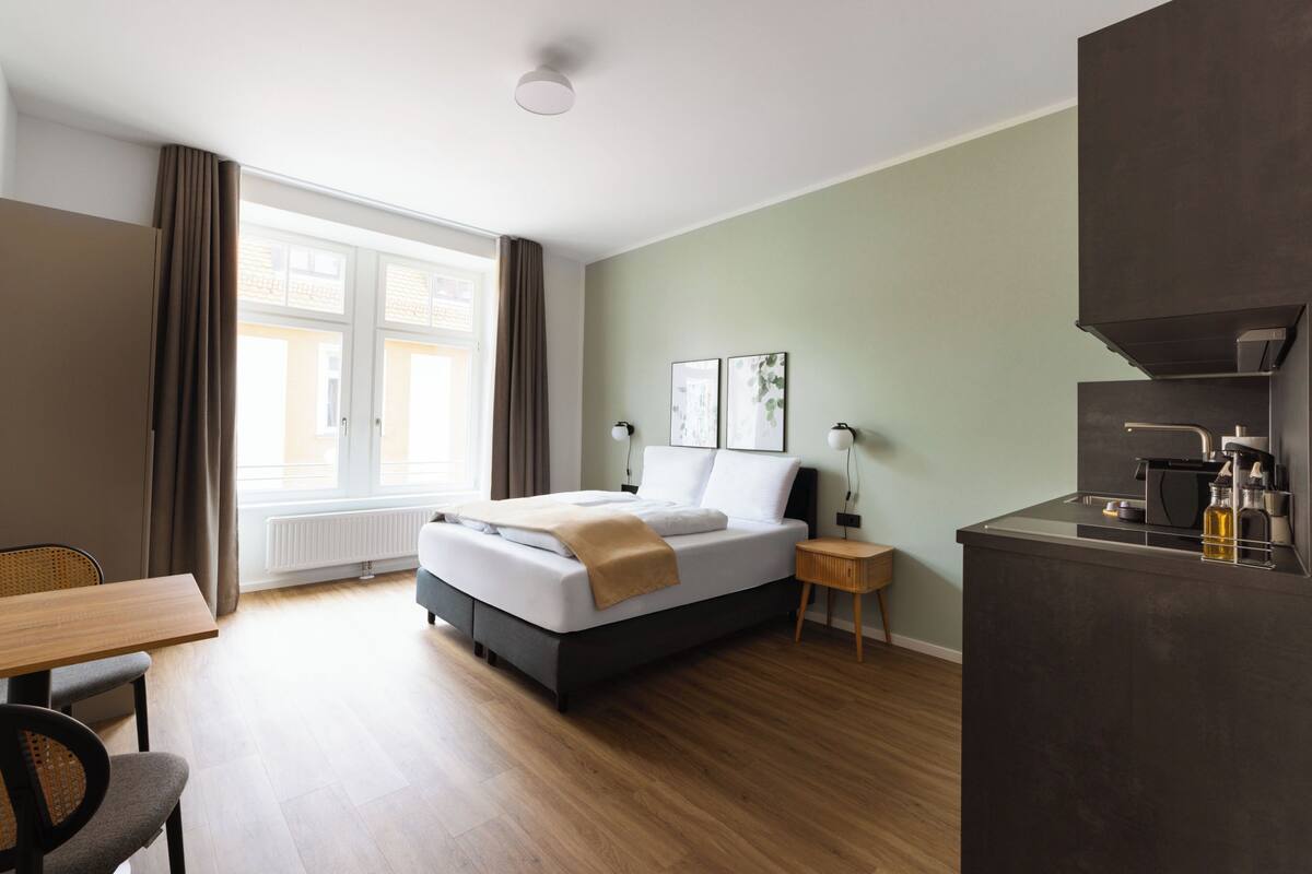Una camera d'albergo con un letto, una scrivania, una sedia, un tavolino, un comodino, una lampada e vista sull'esterno attraverso una finestra.