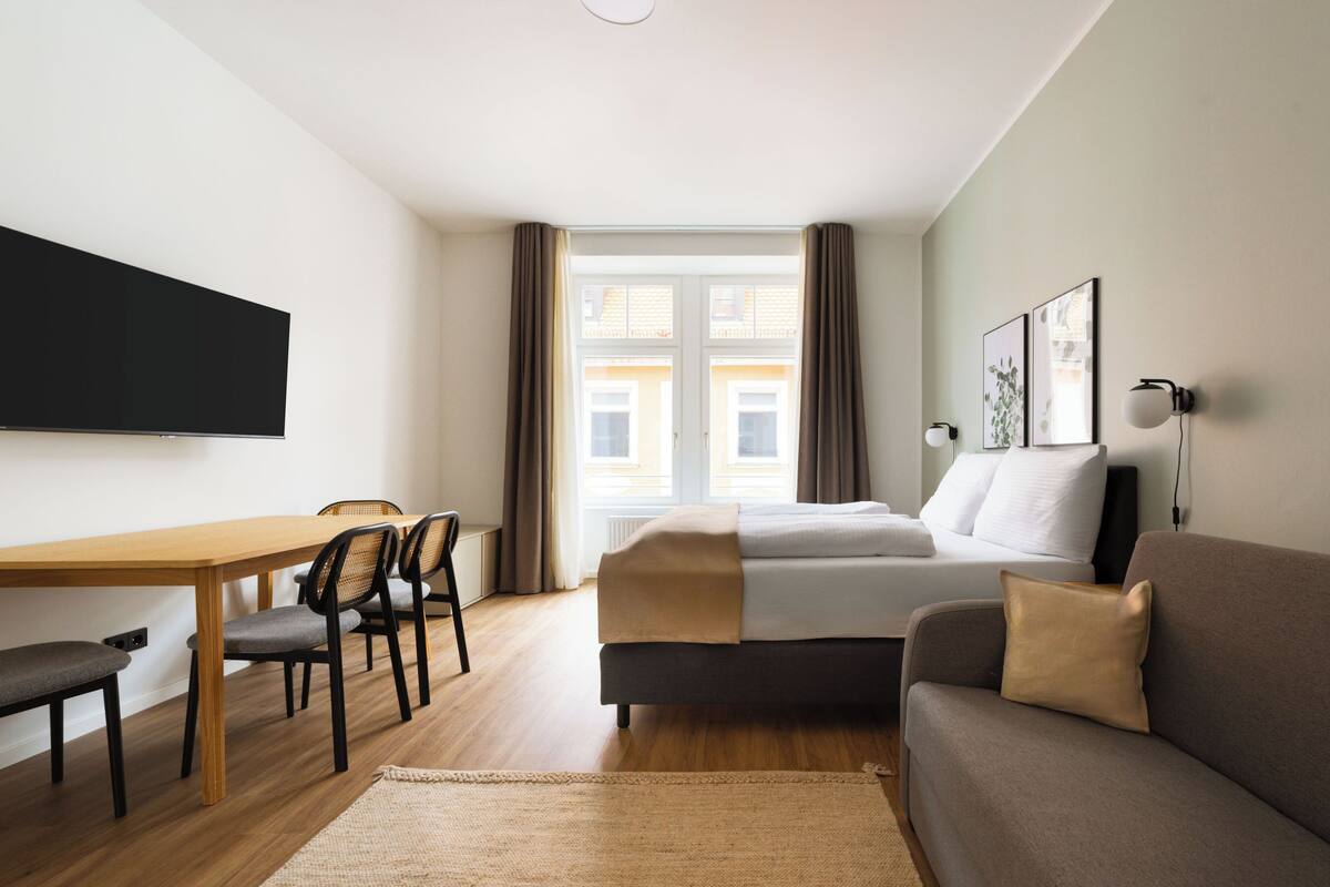 Camera d'albergo con un letto, un divano, una scrivania con sedie, una televisione e una finestra con tende.