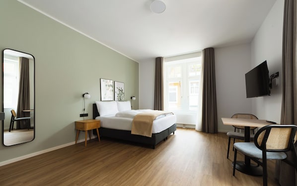 A hotel room with a bed, bedside table, chair, TV, and a window with curtains.
