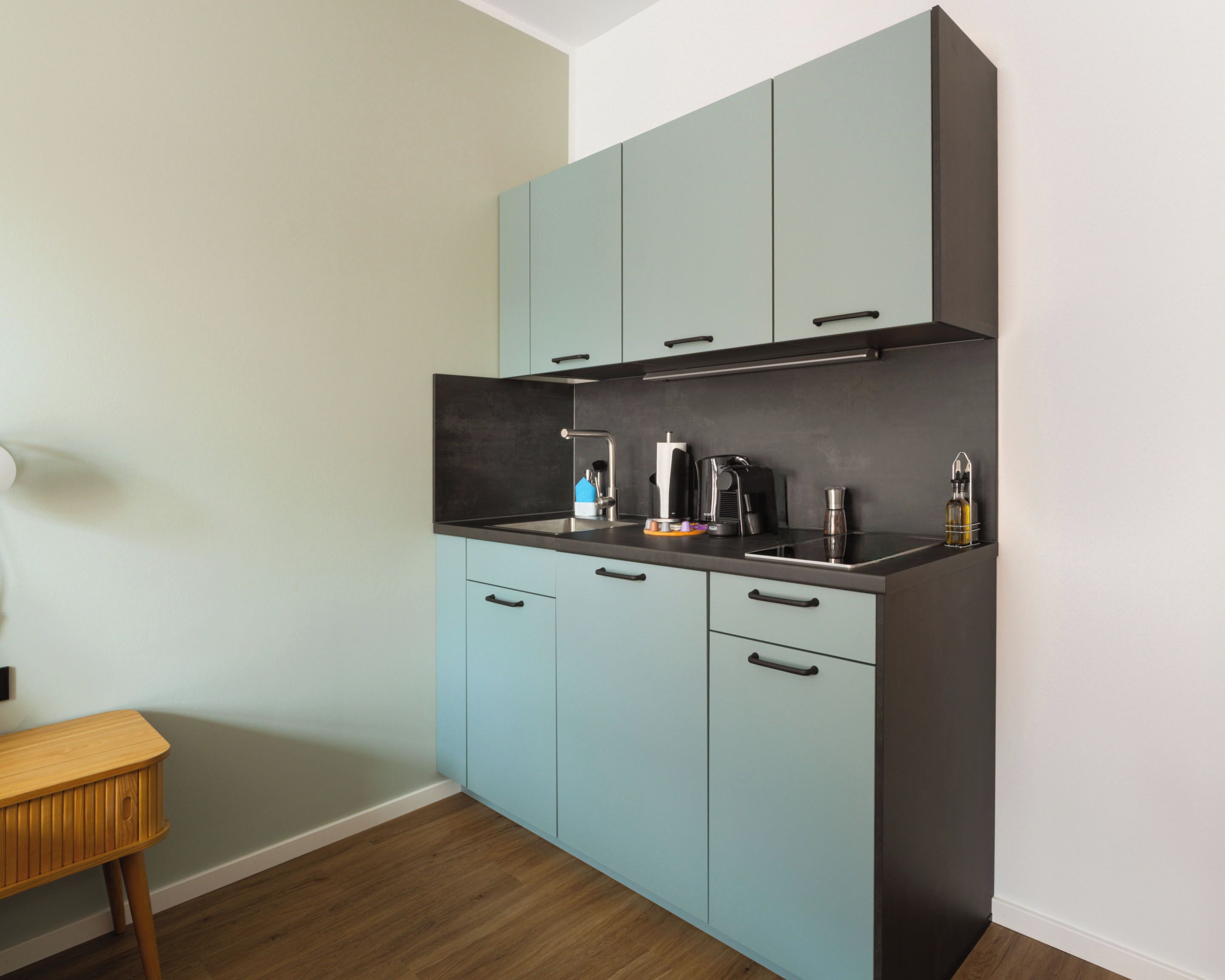Une cuisine de taille réduite avec des meubles de couleur bleu clair, un plan de travail noir et un tabouret en bois.