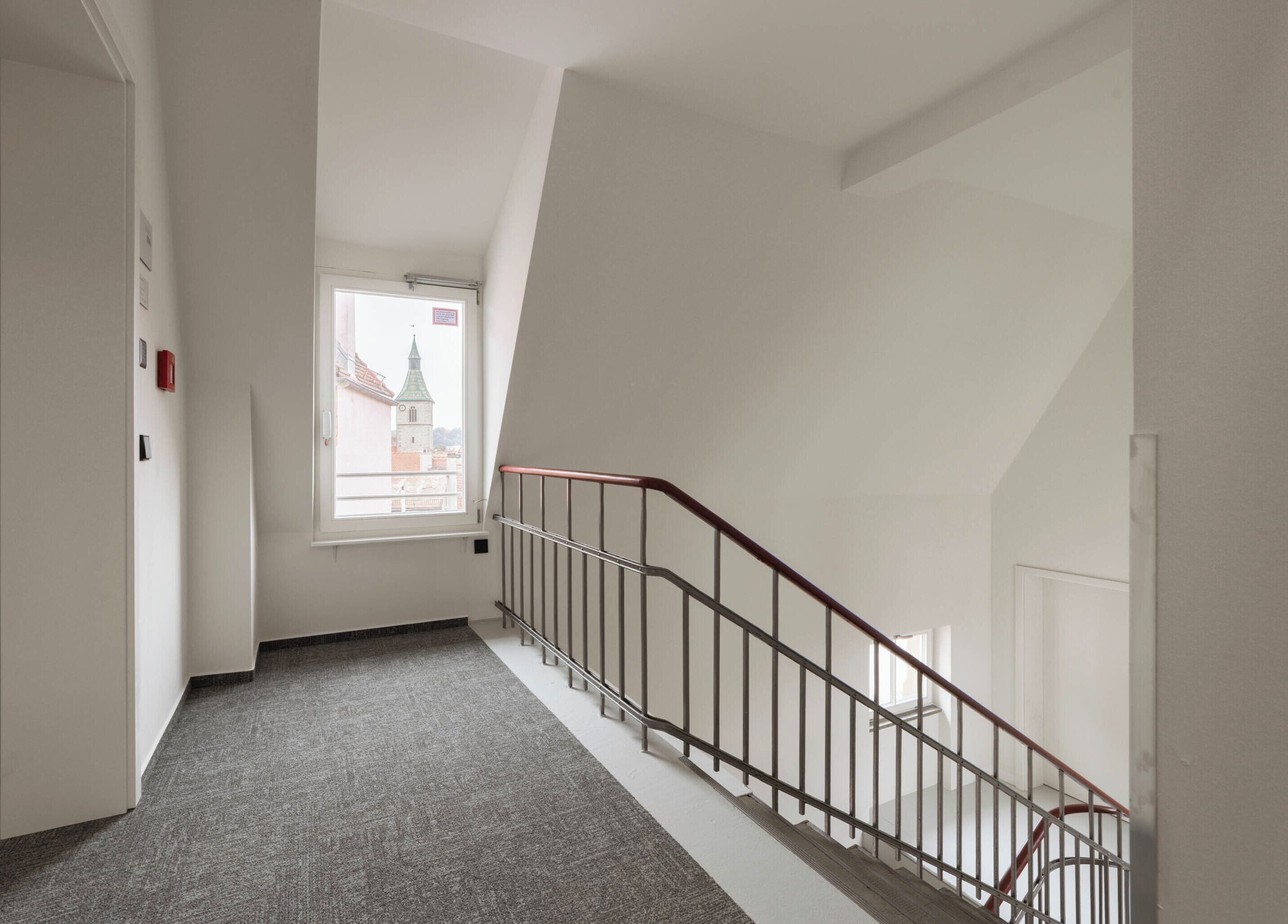 En smal hall med ett heltäckningsmatta på golvet, en trappa med metallräcke och ett fönster med utsikt över en byggnad.