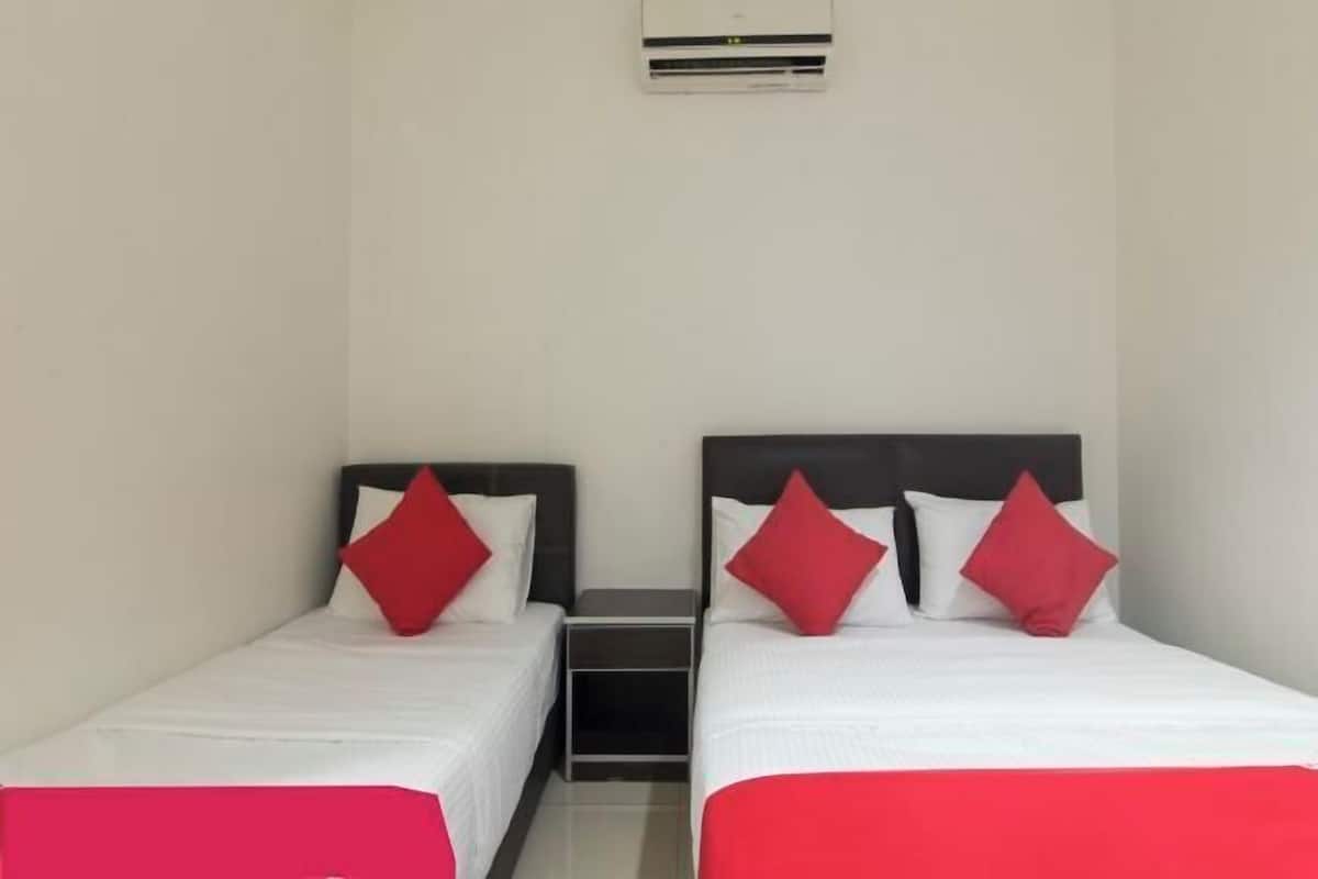 Ein Hotelzimmer mit zwei Einzelbetten, jedes mit einem roten Kopfkissen und einer weißen Bettdecke. Zwischen den Betten befindet sich ein kleiner Nachttisch, und an der Wand darüber ist eine Klimaanlage angebracht.