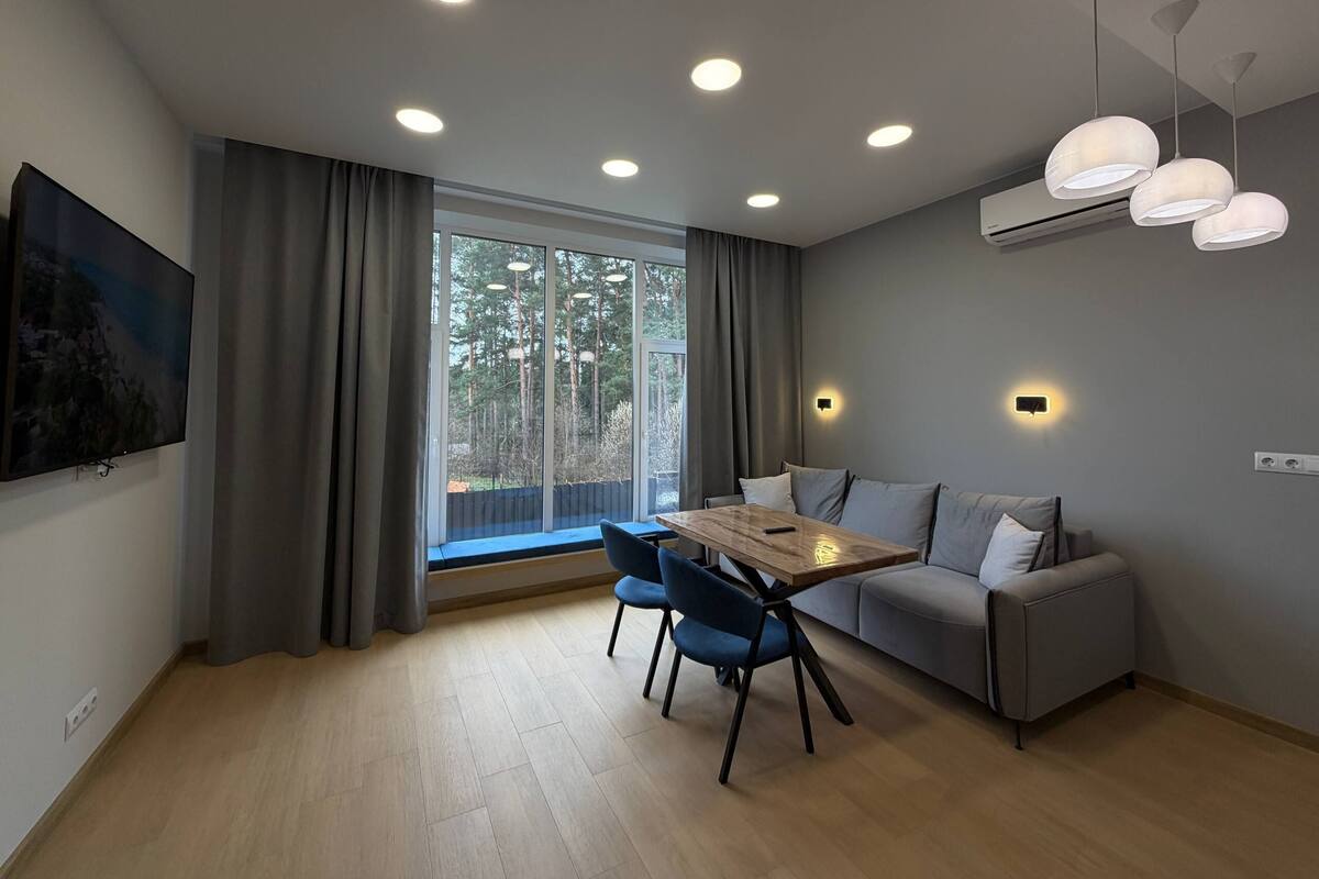 A modern living room with a wooden dining table, blue chairs, a grey sofa, and a large window with curtains.