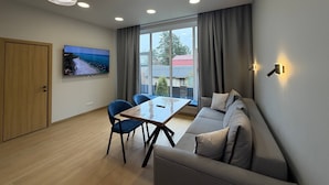 A modern living room with a sofa, a dining table with chairs, a TV mounted on the wall, and a large window with curtains. - ForRest Cozy Apartments Minsk (Minsk)