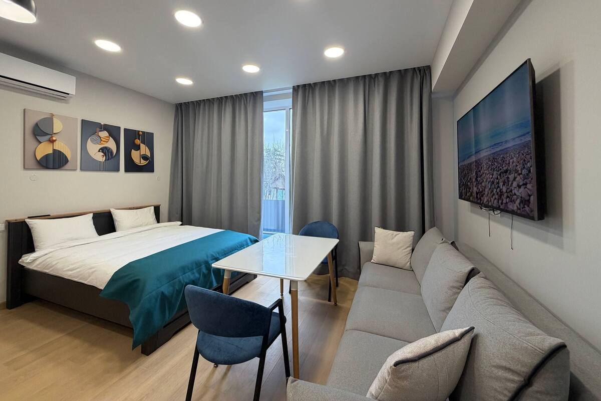 A modern hotel room with a bed, a sofa, a dining table, and a TV.
