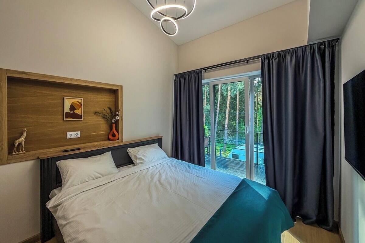A bedroom with a bed, a wooden headboard, a wall-mounted shelf with decorative items, a ceiling light fixture, and a view of a balcony with curtains.