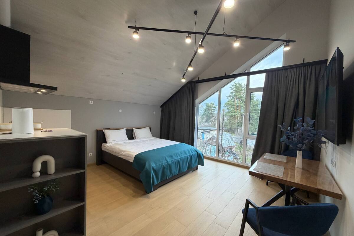 A modern bedroom with a bed, a desk, a chair, a shelf with decorative items, and a large window with curtains.