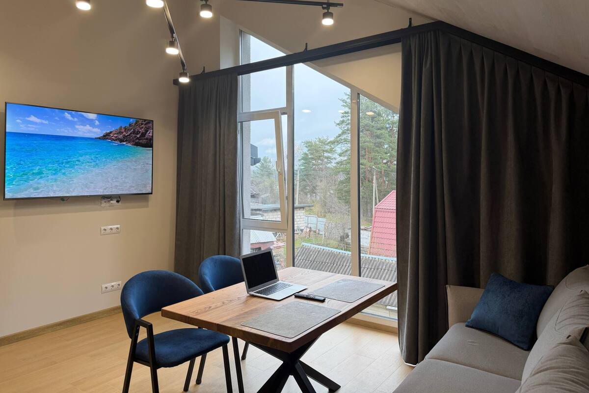 A room with a wooden table, two chairs, a laptop, a TV, and a large window with curtains.