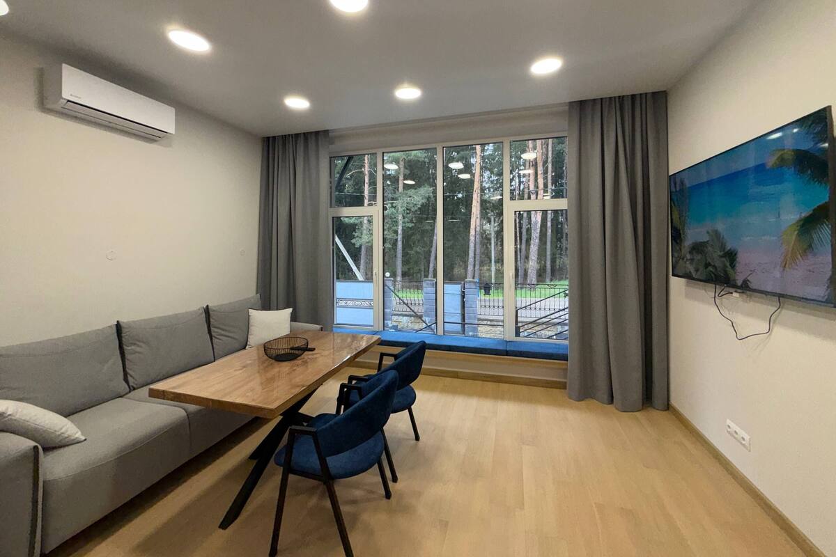 A modern room with a sofa, a dining table with chairs, a large window with curtains, and a mounted TV.