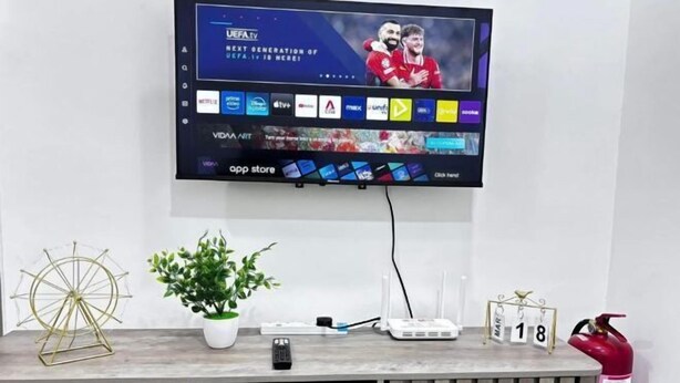 A flat-screen TV mounted on a wall displaying UEFA TV, with a remote control, a plant, a decorative item, and a fire extinguisher on a wooden table below. - JQ PREMIER SUITES T1 L07 (Kota Kinabalu)