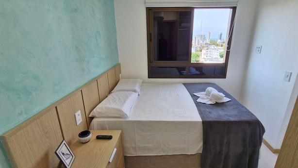A small, neatly arranged bedroom with a single bed, a bedside table with a remote control and a small decorative item, a window with a view of buildings, and a plain wall. - COFDR0104 - Edifício House Federação (Salvador)