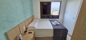 A small, neatly arranged bedroom with a single bed, a bedside table with a remote control and a small decorative item, a window with a view of buildings, and a plain wall. - COFDR0104 - Edifício House Federação (Salvador)