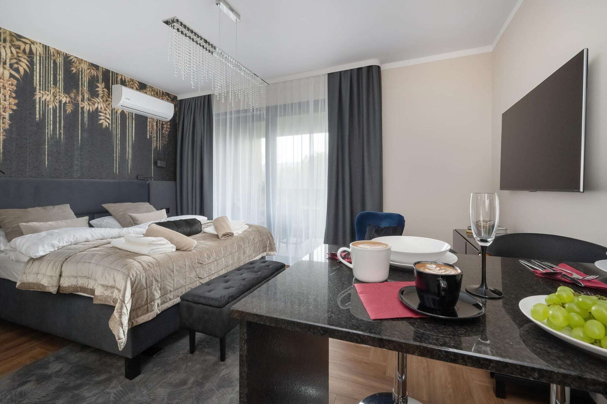 Une chambre d’hôtel moderne équipée d’un lit, d’un bureau avec tout le nécessaire pour préparer du café et d’un coin repas agrémenté d’une assiette de raisins.