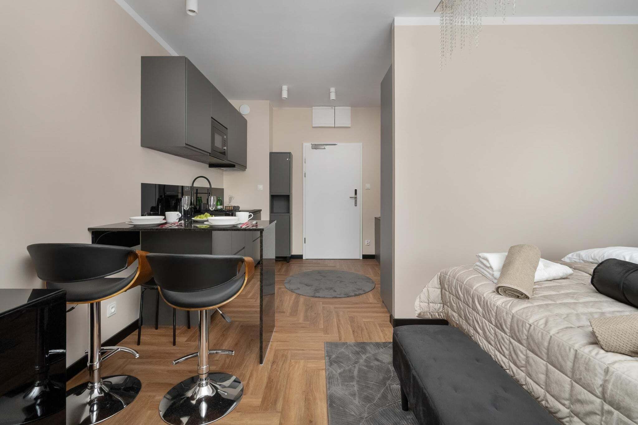 Ein kompaktes Hotelzimmer mit einer Küchenzeile, einem Bett mit grauer Bettdecke und einer kleinen Sitzecke mit zwei Stühlen.