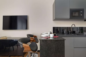 Une cuisine moderne équipée d’un four à micro-ondes encastré, d’un coin repas avec une chaise noire et d’un téléviseur à écran plat fixé au mur.