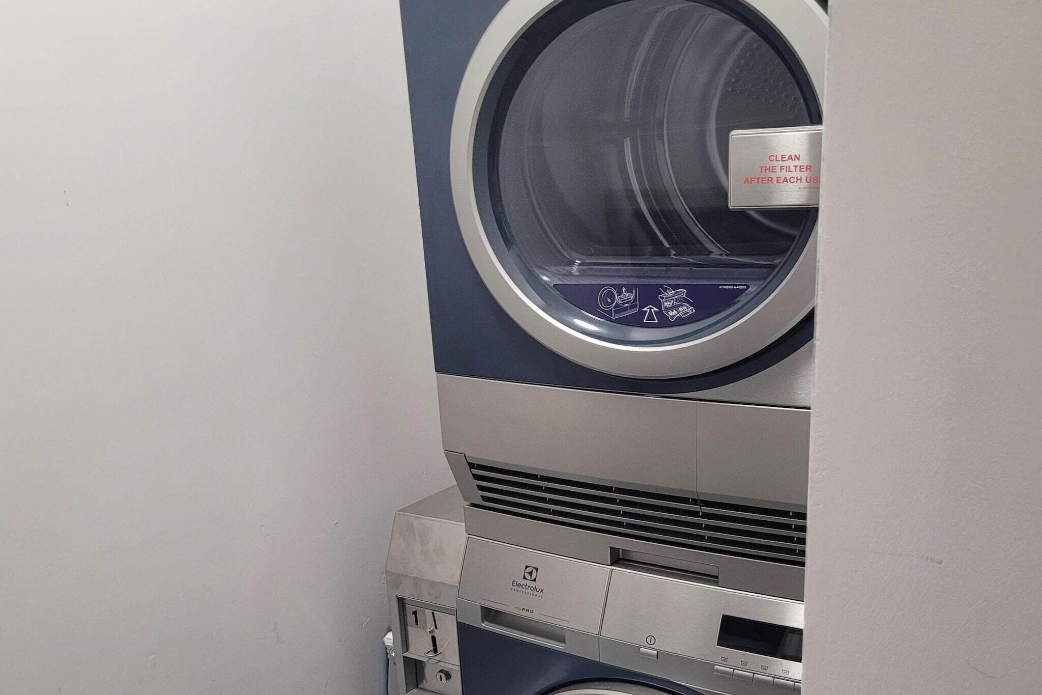 A front view of a washing machine with a front-loading door, a control panel with buttons, and a label instructing to clean the filter after each use.