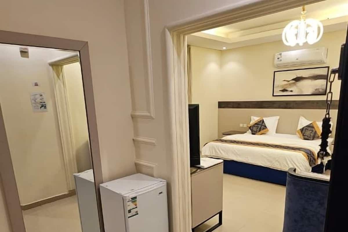 A hotel room with a bed, a desk, a mirror, a small refrigerator, and a ceiling light.