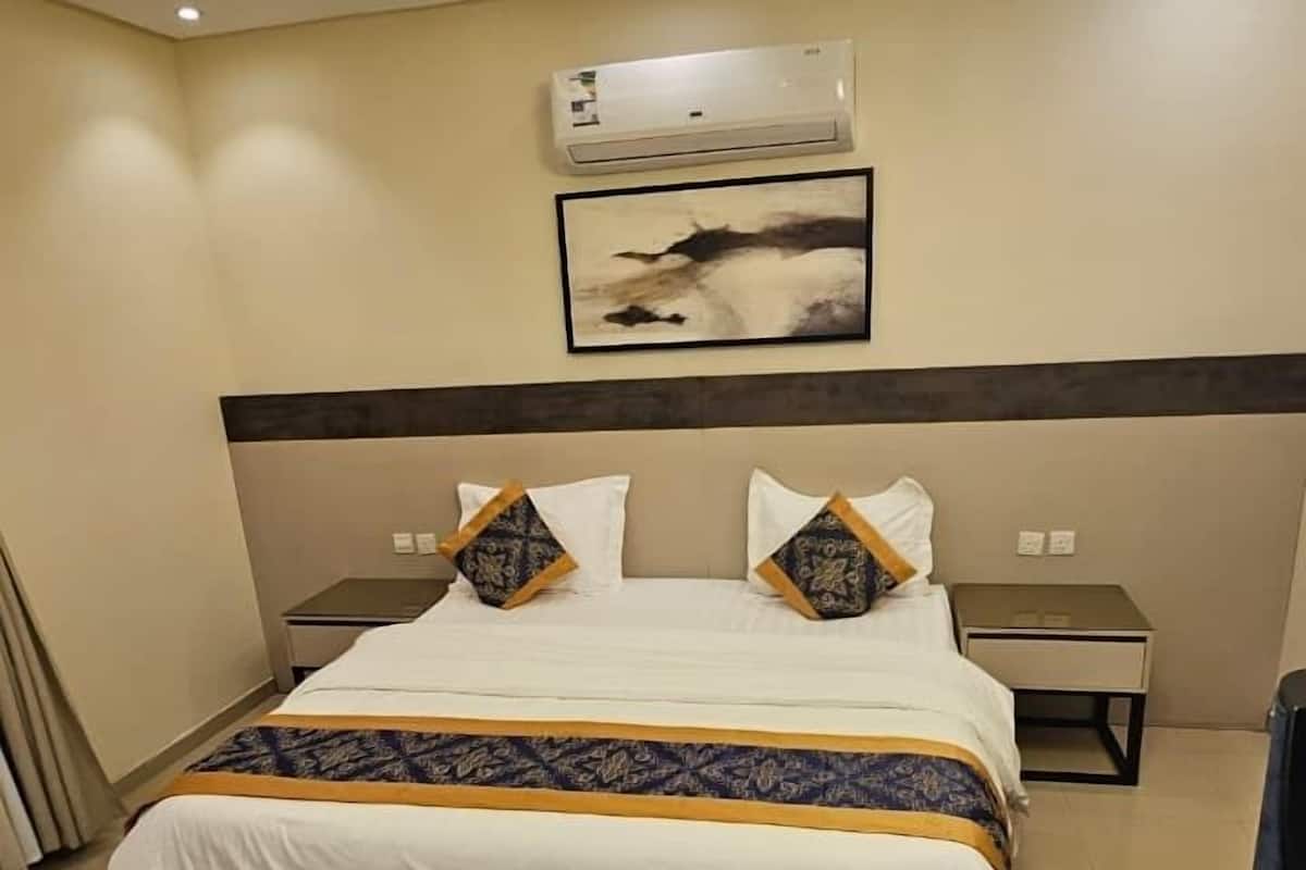 A hotel room with a large bed, a nightstand, a desk, a modern chandelier, and an air conditioning unit on the wall.