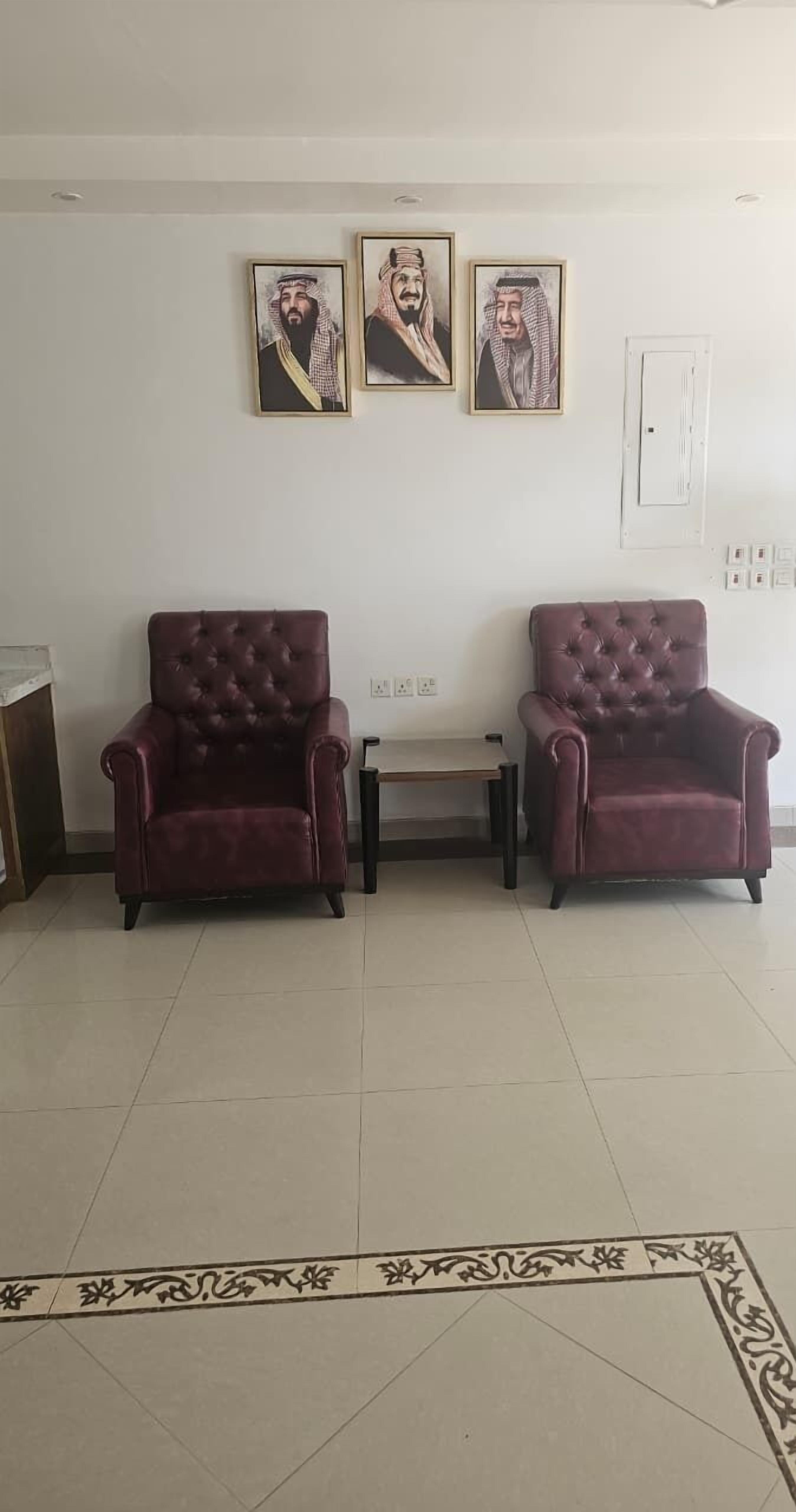 Ein Zimmer mit zwei dunkelroten, gepolsterten Sesseln, die einander zugewandt sind, einem kleinen Tisch dazwischen und drei gerahmten Porträts an der Wand.
