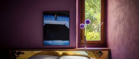 A bedroom with a bed, a window with a view of greenery, and a painting on the wall.