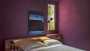 Un dormitorio con cama, una mesita de noche con lámpara, una ventana con vistas a zonas verdes y un cuadro en la pared.