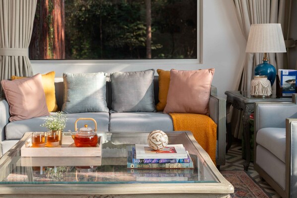 A modern living room with a glass coffee table, a sofa with cushions, a side table with a lamp, and a view of trees through a window.