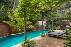 A swimming pool surrounded by lush greenery, with a pergola and a lounge chair.