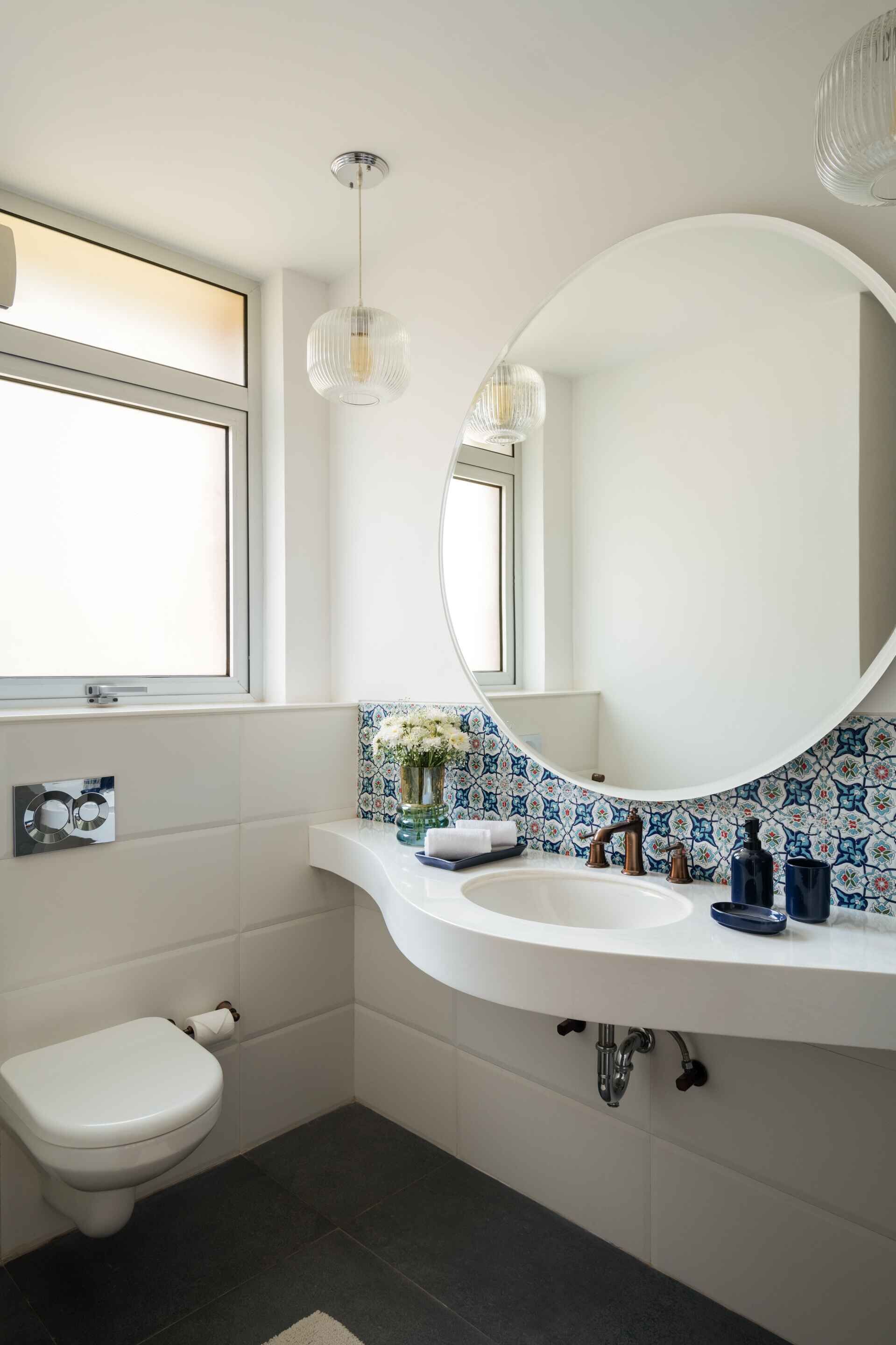 Ein modernes Badezimmer mit einem weißen Waschbecken, einem runden Spiegel, einer Toilette und einem Fenster mit Ausblick.