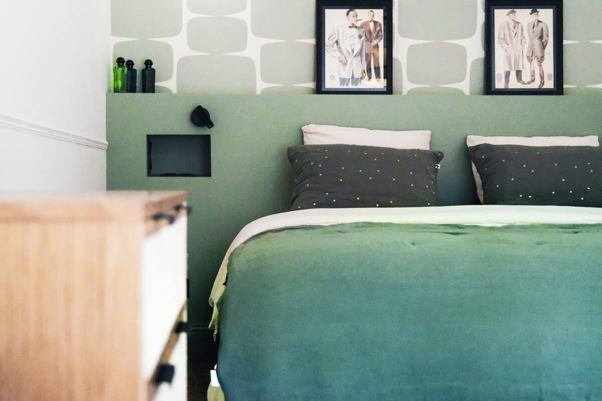 Quarto com colcha verde, duas almofadas com detalhes dourados e duas imagens emolduradas na parede.