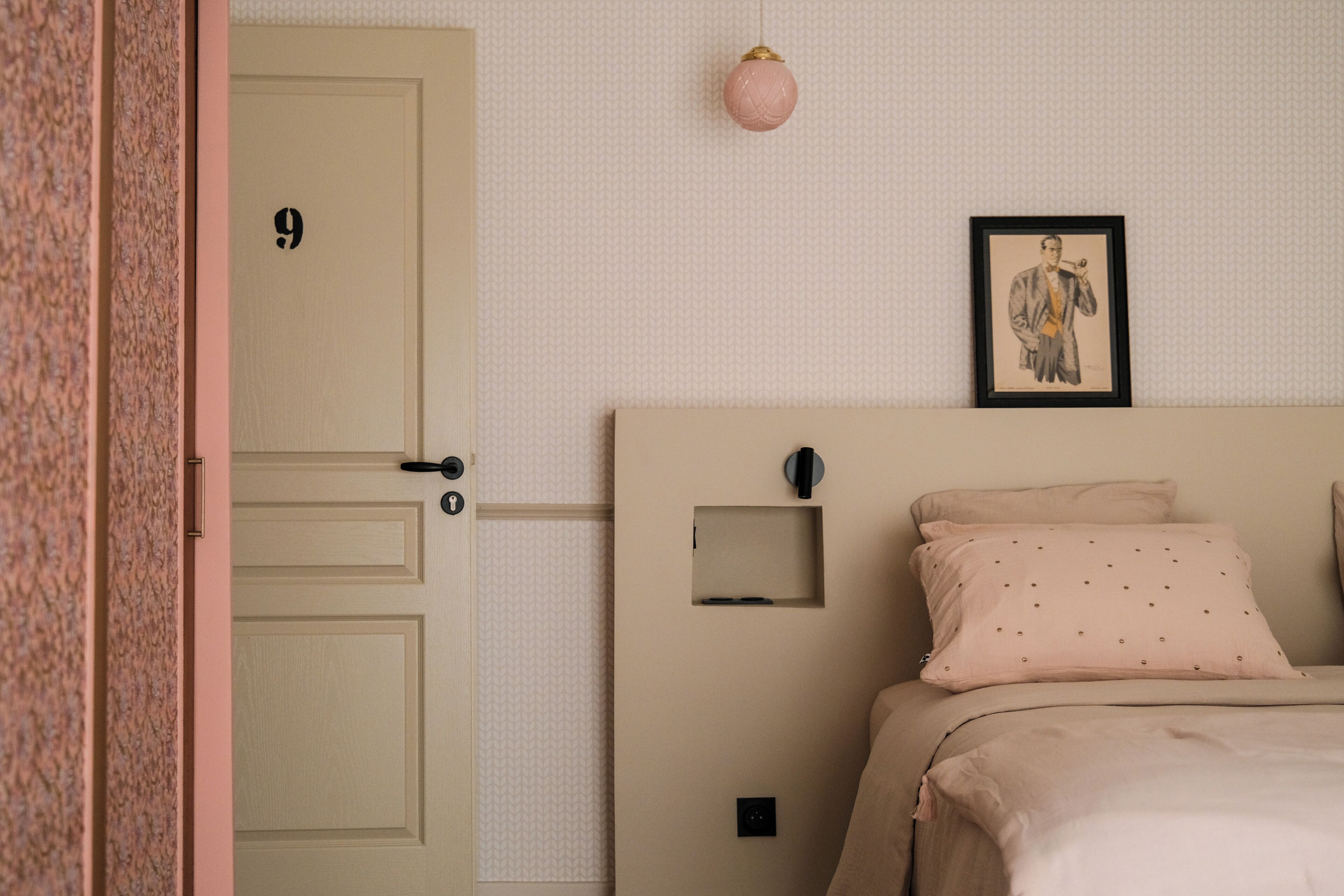A bedroom with a beige door, a framed picture on the wall, a small bedside table, and a bed with pink bedding.
