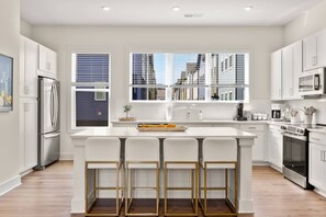 Une cuisine moderne avec des meubles blancs, un îlot central doté de tabourets de bar, des appareils en acier inoxydable et de grandes fenêtres offrant une vue sur la ville.