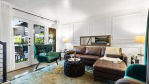 Una sala de estar con un sofá de cuero marrón, un sillón verde y un reposapiés negro. Hay una mesa de vidrio con una botella y un vaso encima.