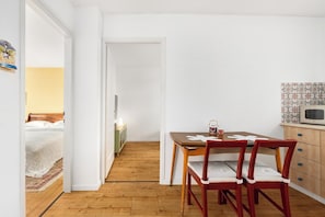 A compact room with a wooden floor, a dining table with chairs, a bed with a white duvet, a small cabinet, and a wall-mounted microwave.
