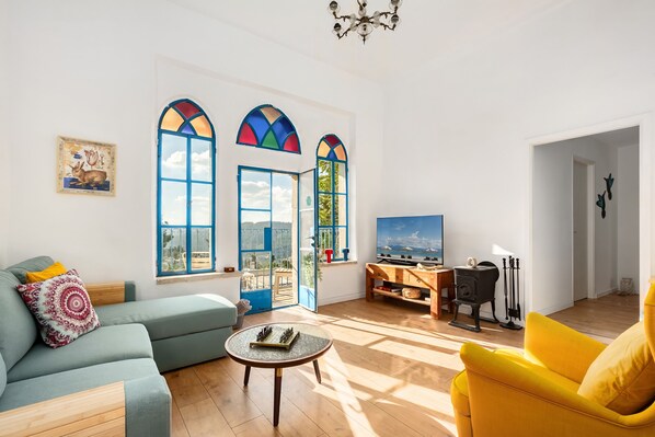 Ein Wohnzimmer mit einer Couch, einem Sessel und einem Couchtisch. Zwei Fenster mit farbigem Buntglas, ein Kamin und ein Fernseher sind ebenfalls vorhanden.