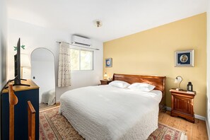 Ein Schlafzimmer mit einem großen Bett, einem Nachttisch aus Holz, einem Schreibtisch mit Lampe und einem Fenster mit Vorhängen.