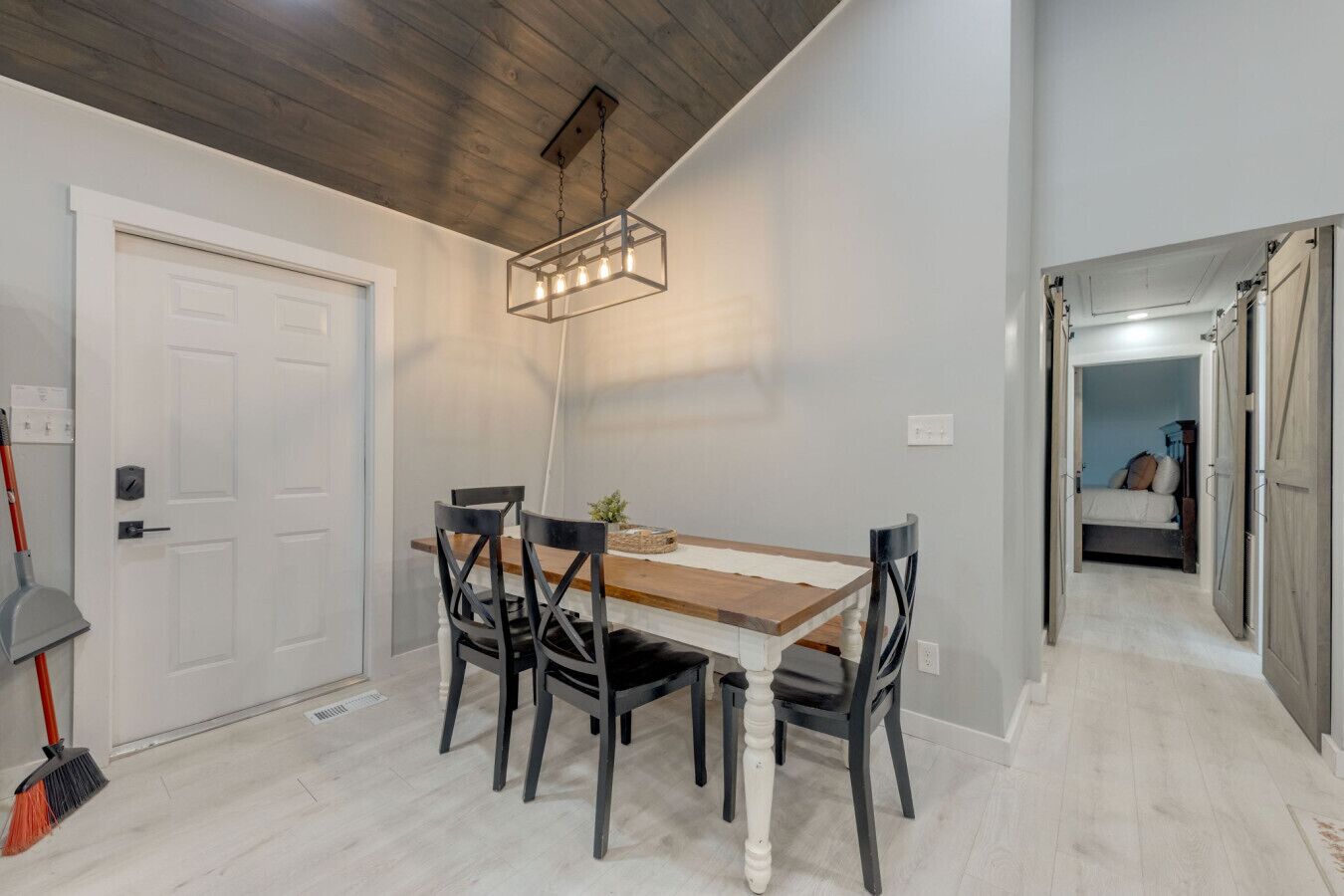 A dining area with a wooden table and four chairs, a hanging light fixture, and a door with a broom leaning against it.