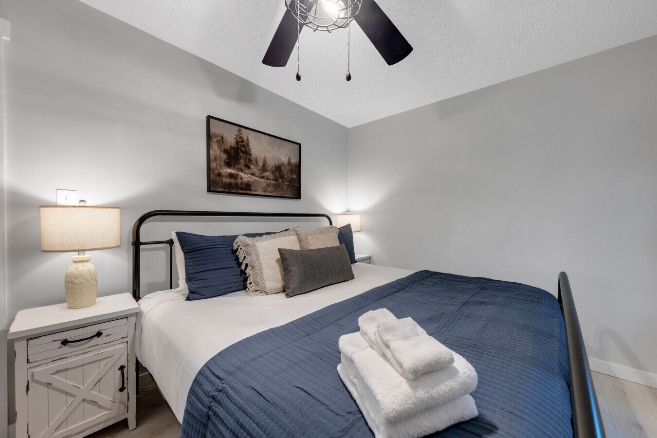 A bedroom with a bed, a nightstand, a lamp, a ceiling fan, and a painting on the wall.