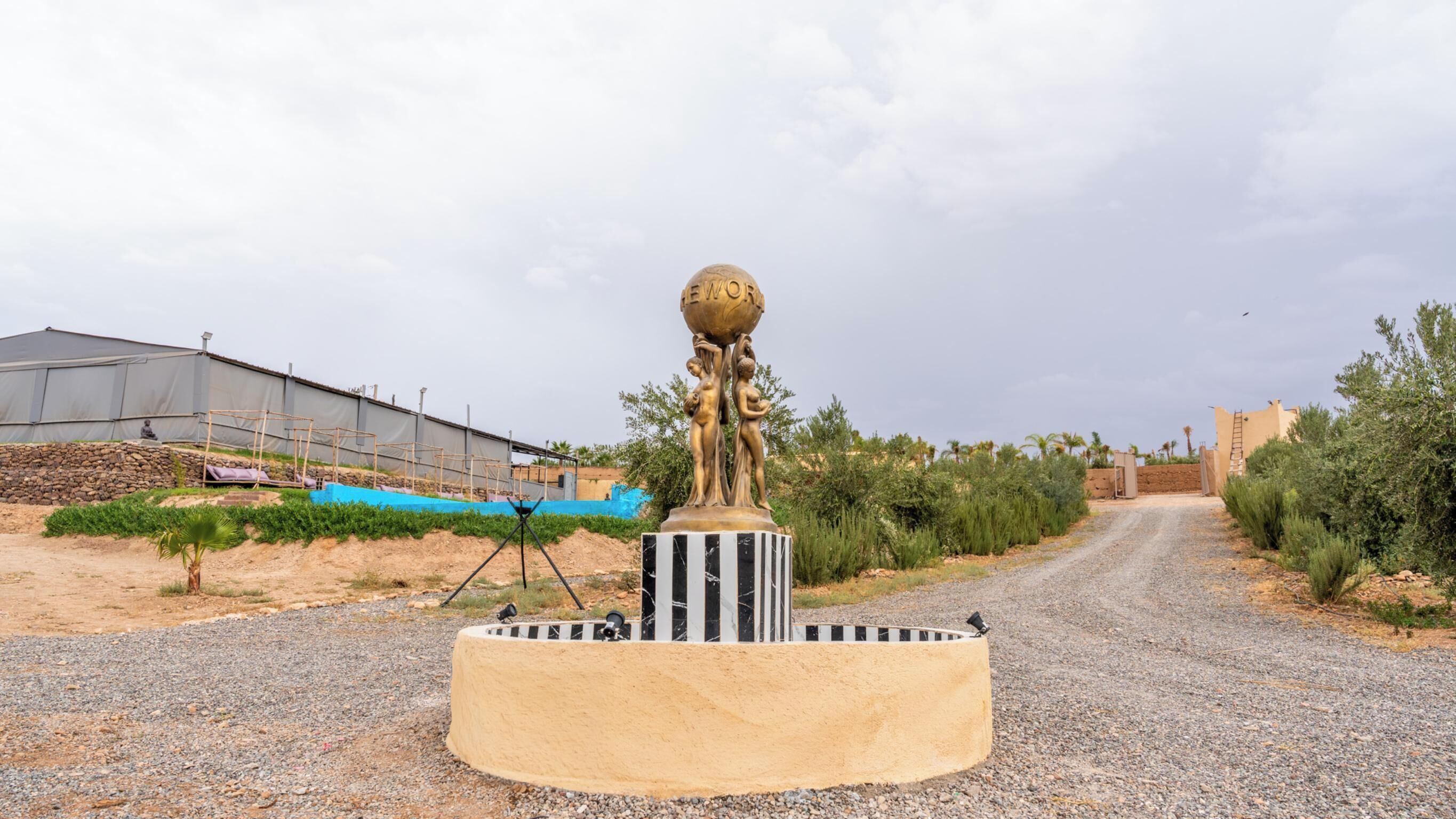 A sculpture on a pedestal in a rural setting with a gravel path, agricultural fields, and a building in the background.