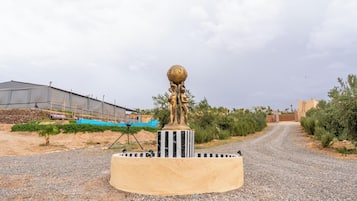 A sculpture on a pedestal in a rural setting with a gravel path, agricultural fields, and a building in the background.