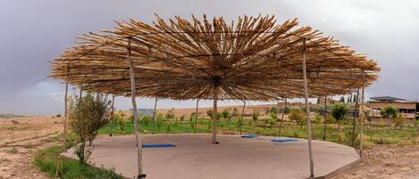 A circular outdoor area with a thatched roof structure, surrounded by a concrete floor, and a few scattered trees.