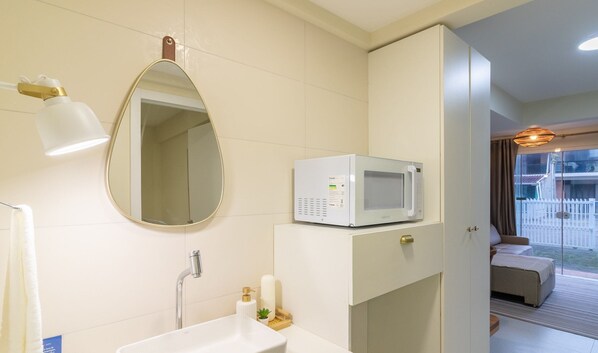 Une salle de bain moderne avec un lavabo blanc, un miroir, une serviette, une douche et un four à micro-ondes posé sur un meuble.